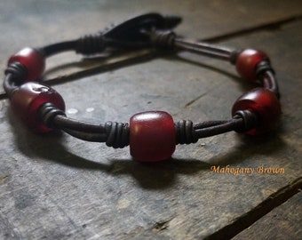 May include: A leather bracelet with red glass beads and a brown leather closure. The bracelet is on a wooden surface. "Mahogany Brown" is written on the surface.