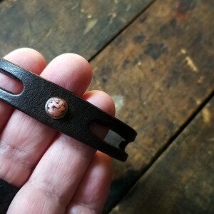 Puede incluir: Un brazalete de cuero marr&oacute;n con un acento de piedra rosa y blanca.