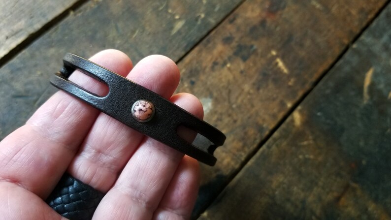 Puede incluir: Un brazalete de cuero marr&oacute;n con un acento de piedra rosa y blanca.