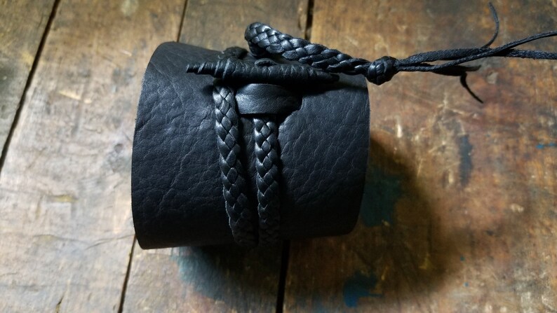 May include: A black leather cuff bracelet with a braided leather accent. The cuff is thick and wraps around the wrist. A braided leather cord and knot detail the top of the cuff.