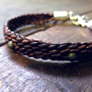 Puede incluir: Un brazalete de cuero marr&oacute;n con un dise&ntilde;o trenzado y detalles plateados. El brazalete tiene peque&ntilde;as cuentas met&aacute;licas espaciadas a lo largo del cuero trenzado.