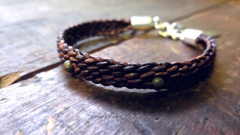Puede incluir: Un brazalete de cuero marr&oacute;n con un dise&ntilde;o trenzado y detalles plateados. El brazalete tiene peque&ntilde;as cuentas met&aacute;licas espaciadas a lo largo del cuero trenzado.