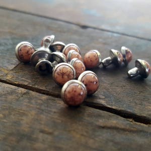 May include: A close-up of a pile of small, round, pink and brown decorative studs with silver metal bases. The studs have a marbled appearance.