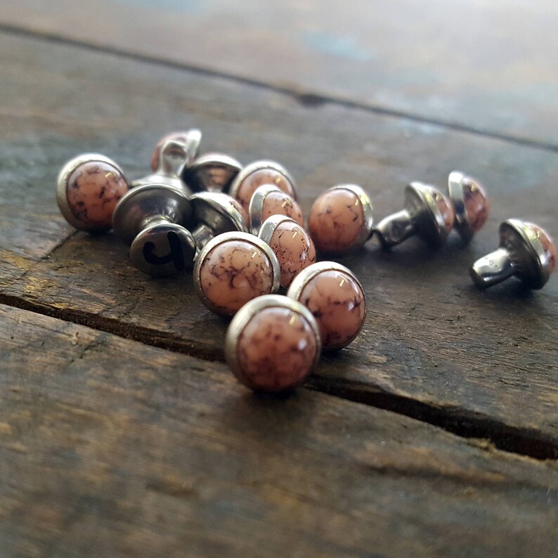 May include: A close-up of a pile of small, round, pink and brown decorative studs with silver metal bases. The studs have a marbled appearance.