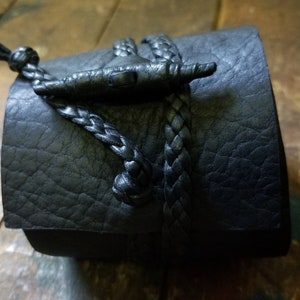 May include: Black leather cuff bracelet with a braided leather closure. The bracelet features a textured surface and a decorative toggle clasp. The cuff is a dark, neutral color, suitable for various styles.