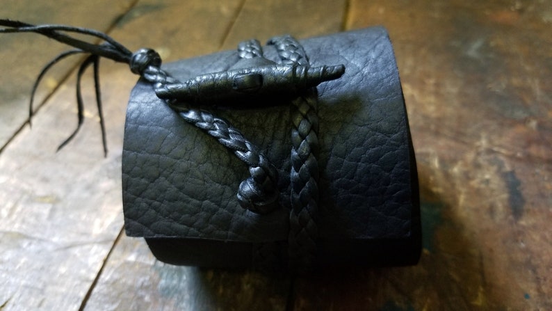 May include: Black leather cuff bracelet with a braided leather closure. The bracelet features a textured surface and a decorative toggle clasp. The cuff is a dark, neutral color, suitable for various styles.