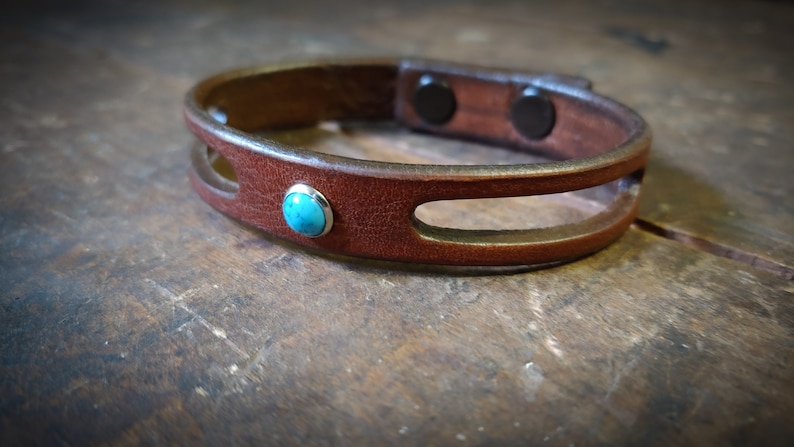 Puede incluir: Un brazalete de cuero marr&oacute;n con una piedra turquesa y un cierre magn&eacute;tico.