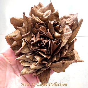 May include: A bronze-colored leather flower accessory featuring multiple layered petals arranged in a bloom shape. The text "New 15-Layer Collection" is printed in a gold script font at the bottom of the image.