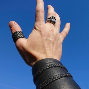 May include: A hand wearing a black braided leather ring, a silver ring with a textured design, and a black leather wrist cuff with braided accents. The background is a clear blue sky.