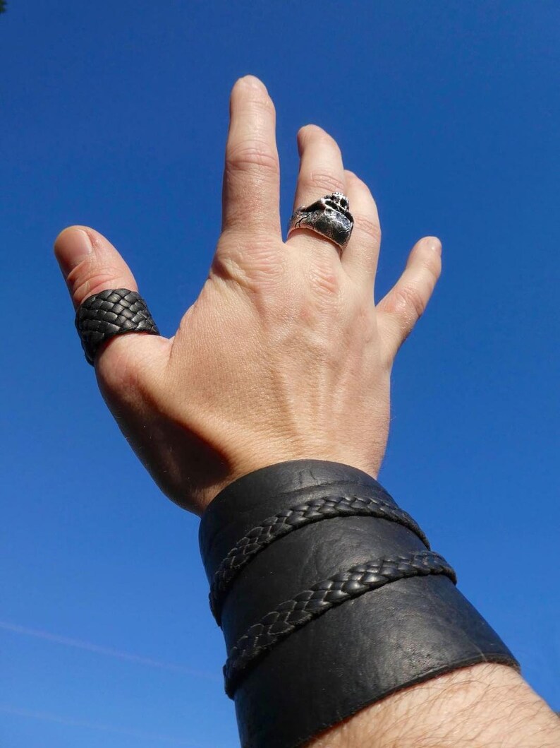May include: A hand wearing a black braided leather ring, a silver ring with a textured design, and a black leather wrist cuff with braided accents. The background is a clear blue sky.