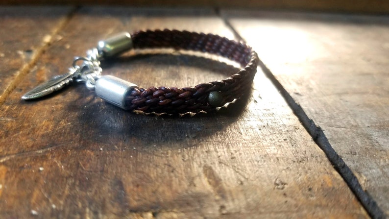 Puede incluir: Un brazalete de cuero marr&oacute;n con un dije de pluma de plata y dos acentos de metal plateado. El brazalete est&aacute; trenzado y tiene una cuenta marr&oacute;n oscura.