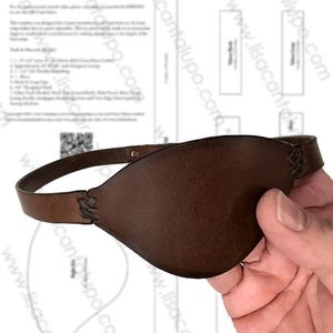 Könnte beinhalten: Eine braune Leder-Augenklappe mit einem Riemen. Die Augenklappe wird von einer Hand gehalten, und das Design umfasst eine gebogene Form, die sich dem Auge anpasst. Der Riemen ist mit einem geflochtenen Design befestigt. Der Hintergrund enthält eine Designvorlage.