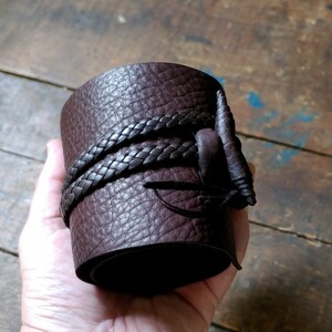 May include: A wide, dark brown leather cuff bracelet with a textured surface. The bracelet is wrapped with a braided leather strap and features a decorative leather accent. The cuff is approximately 3 inches wide.