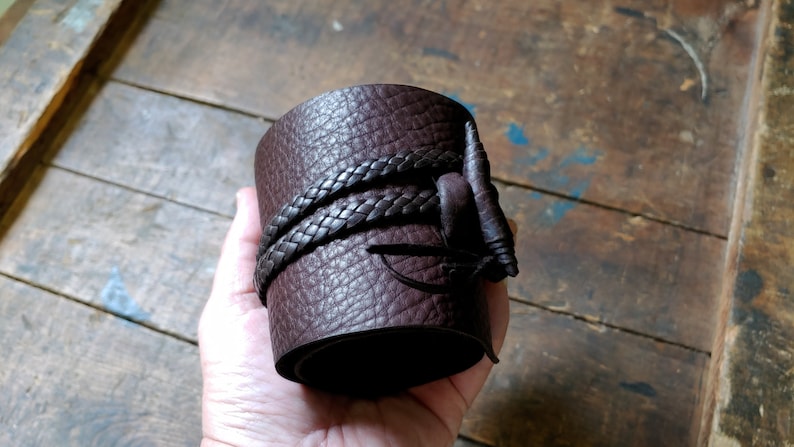 May include: A wide, dark brown leather cuff bracelet with a textured surface. The bracelet is wrapped with a braided leather strap and features a decorative leather accent. The cuff is approximately 3 inches wide.