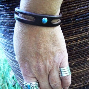 Puede incluir: Un brazalete de cuero marr&oacute;n con un acento de piedra turquesa. El brazalete se lleva en la mu&ntilde;eca de una persona. La persona tambi&eacute;n lleva un anillo de plata con una piedra turquesa en su dedo.