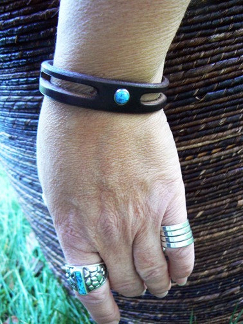 Puede incluir: Un brazalete de cuero marr&oacute;n con un acento de piedra turquesa. El brazalete se lleva en la mu&ntilde;eca de una persona. La persona tambi&eacute;n lleva un anillo de plata con una piedra turquesa en su dedo.