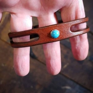 Puede incluir: Un brazalete de cuero marr&oacute;n con un acento de piedra turquesa. El brazalete tiene dos recortes en la correa de cuero.