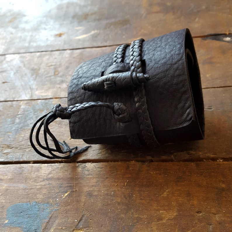 May include: A dark brown leather wrist cuff with a braided strap and toggle closure. The cuff is wrapped around the wrist and secured with a braided leather strap and a decorative toggle. The cuff has a textured surface and a knotted tassel.