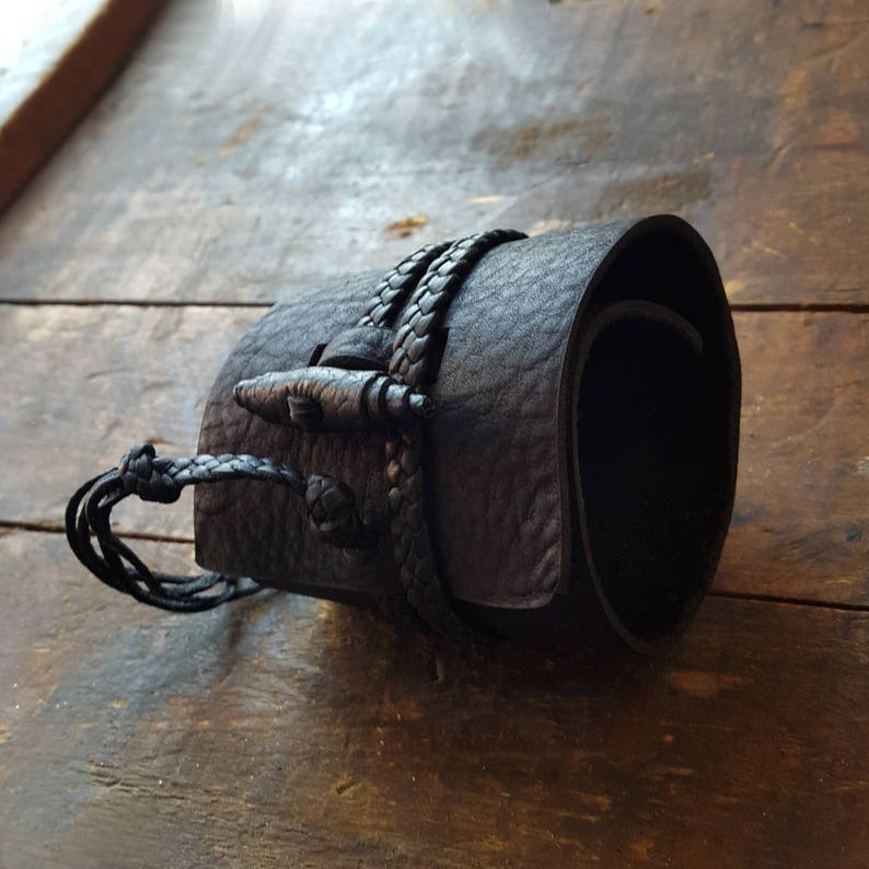 May include: A black leather cuff bracelet with a braided strap and toggle closure. The cuff is rolled and has a textured surface. The bracelet is shown on a wooden surface.