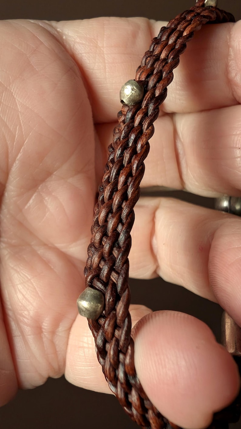 Puede incluir: Un brazalete de cuero marr&oacute;n con detalles de metal plateado. El brazalete est&aacute; tejido en un patr&oacute;n trenzado.