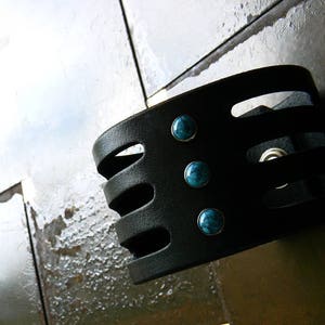 May include: Black leather bracelet with four turquoise stones. The bracelet has a geometric design with cutouts.