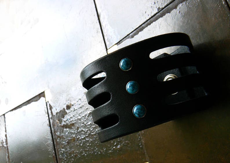 May include: Black leather bracelet with four turquoise stones. The bracelet has a geometric design with cutouts.