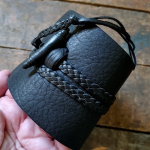 May include: Black leather cuff bracelet with a braided leather accent. The cuff features a unique closure with a leather toggle and loop. The bracelet is a wide style, suitable for a bold statement piece.