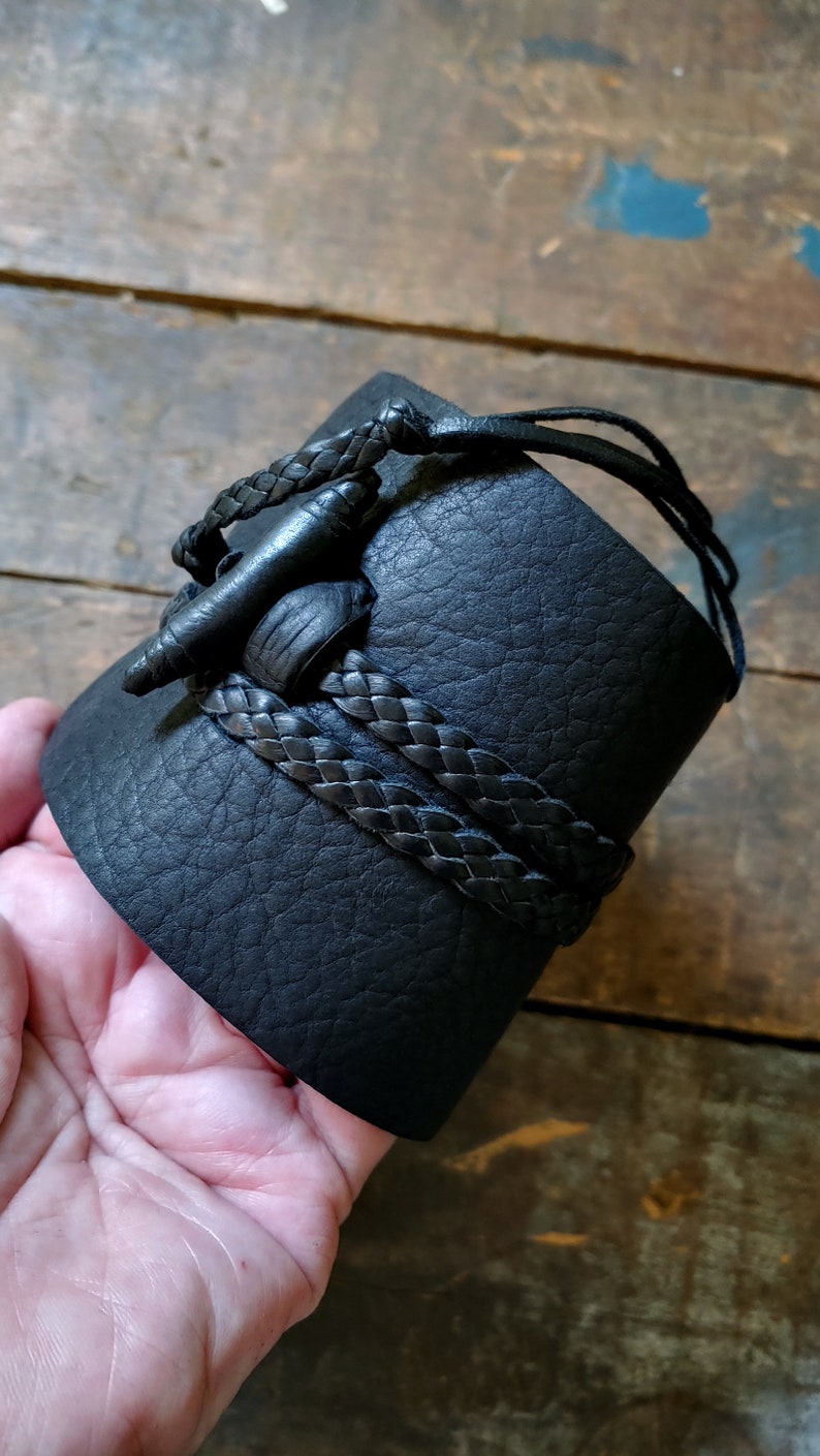 May include: Black leather cuff bracelet with a braided leather accent. The cuff features a unique closure with a leather toggle and loop. The bracelet is a wide style, suitable for a bold statement piece.