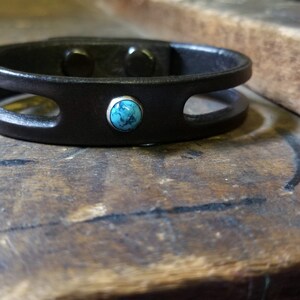 Puede incluir: Un brazalete de cuero negro con un acento de piedra turquesa. El brazalete tiene dos recortes y un dise&ntilde;o simple.