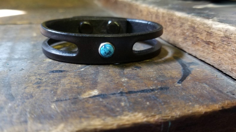 Puede incluir: Un brazalete de cuero negro con un acento de piedra turquesa. El brazalete tiene dos recortes y un dise&ntilde;o simple.