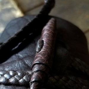May include: Close-up of a dark brown leather sandal with a braided strap and a decorative, cylindrical leather element. The cylindrical element has a spiral design and a small, textured detail.