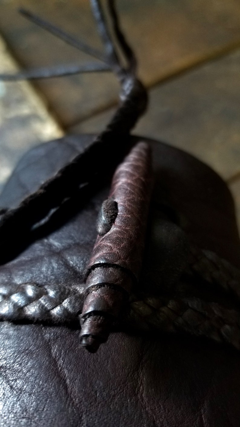 May include: Close-up of a dark brown leather sandal with a braided strap and a decorative, cylindrical leather element. The cylindrical element has a spiral design and a small, textured detail.