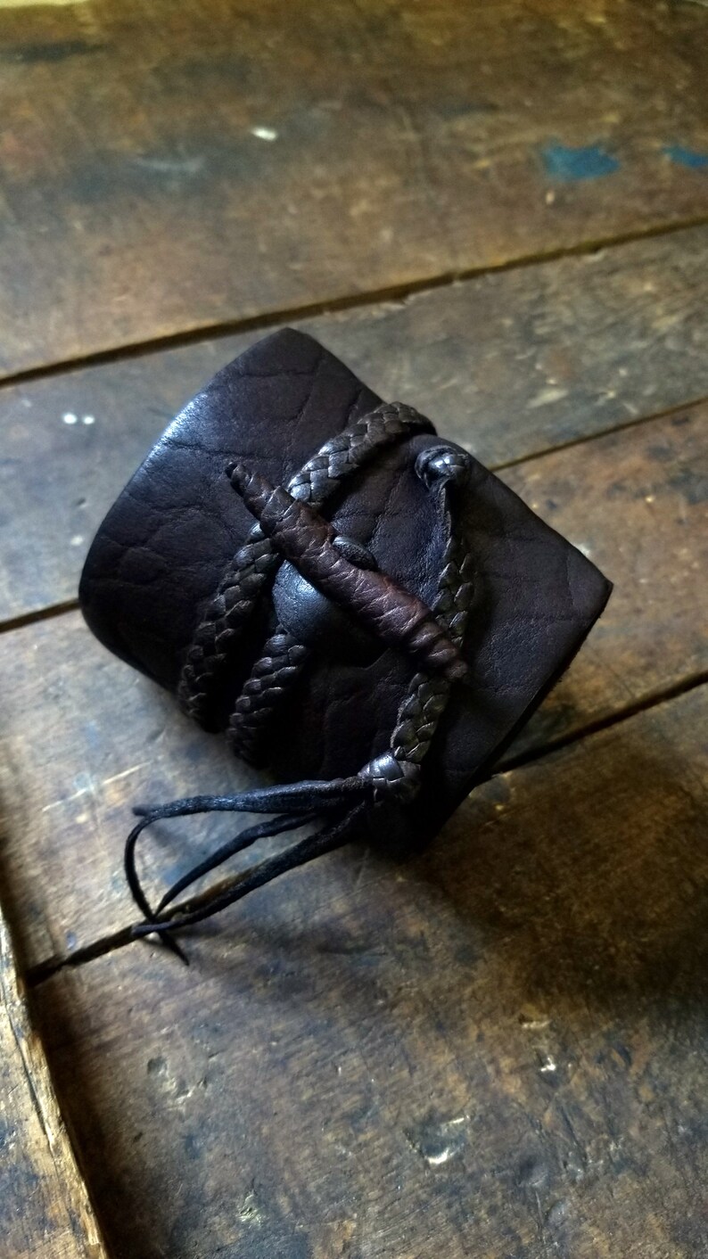 May include: Dark brown leather cuff bracelet with braided leather accents and a central decorative element. The cuff is approximately 2 inches wide and features a textured surface, with a braided leather strap.