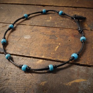 Pode incluir: Um colar de couro preto com contas azuis turquesa. O colar é amarrado com um nó e tem um pequeno fecho de metal.