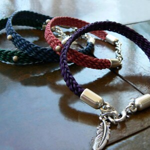 Puede incluir: Tres pulseras trenzadas con detalles plateados. Las pulseras son verde, roja y morada. La pulsera morada tiene un dije de pluma plateada.