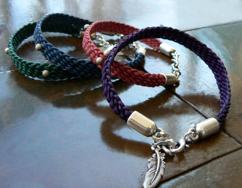 Puede incluir: Tres pulseras trenzadas con detalles plateados. Las pulseras son verde, roja y morada. La pulsera morada tiene un dije de pluma plateada.