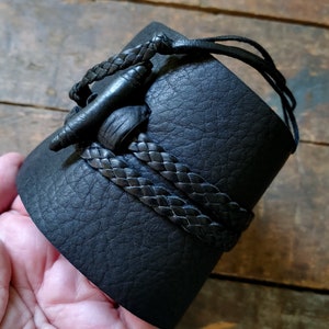 May include: Black leather cuff bracelet with a braided leather accent. The cuff features a unique closure with a leather toggle and loop. The bracelet is a wide style, suitable for a bold statement piece.