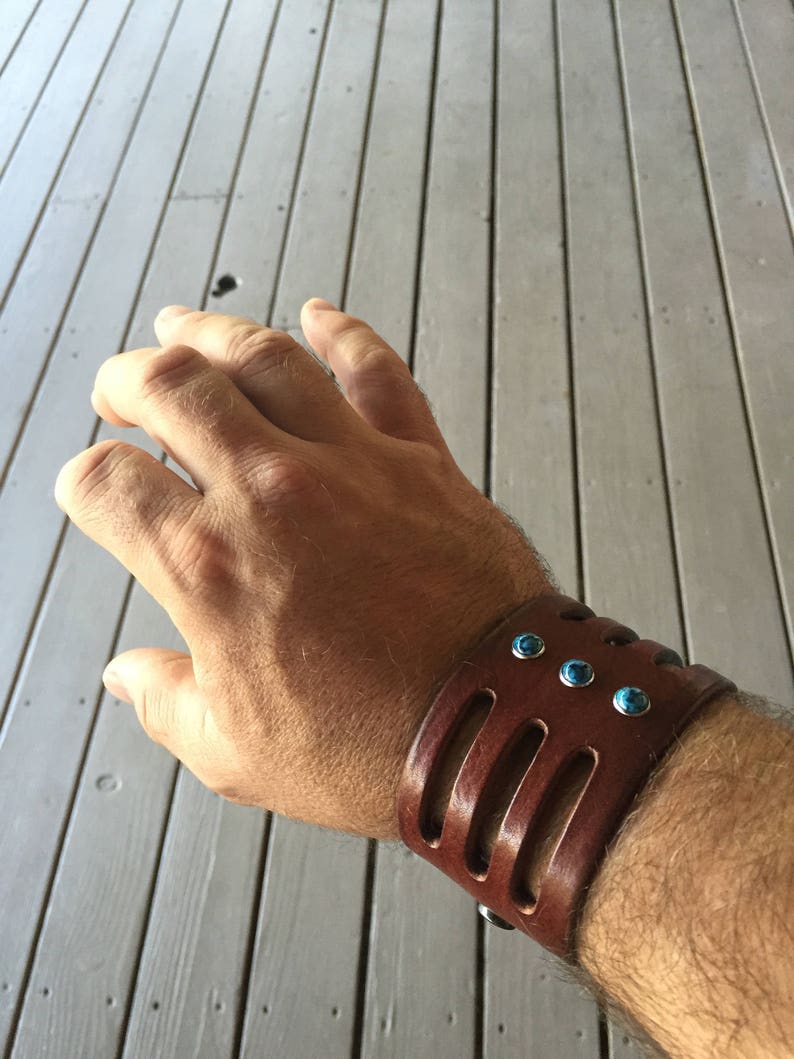 May include: A brown leather cuff bracelet with turquoise stones. The bracelet has three cutouts and is worn on a person's wrist.