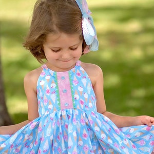 May include: A young girl wearing a blue and pink ice cream cone print dress with a light blue bow in her hair.
