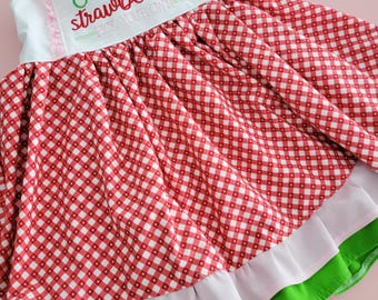 May include: A red and white gingham dress with a white bodice and a green and pink ruffle trim. The bodice has embroidered text that reads "Farm Fresh Strawberries Pick Your Own".