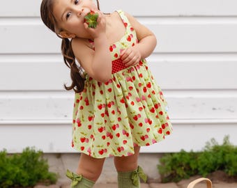 May include: A young girl wearing a green and red strawberry print dress, red cowboy boots, and green socks with bows, eats a strawberry while standing on a brick patio. A wicker basket filled with strawberries sits on the patio.