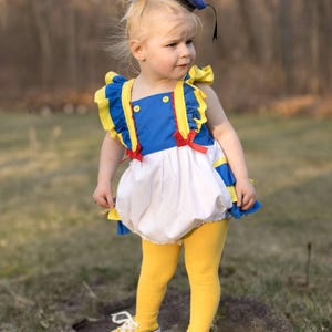 May include: A toddler in a Donald Duck costume, featuring a blue and white top with yellow accents, a white bubble skirt, and yellow leggings. The outfit is completed with a blue sailor hat and yellow sneakers.