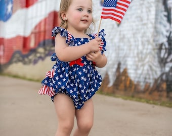 May include: A young child wearing a blue and white star-patterned dress with red accents holds a small American flag in front of a mural of an eagle and an American flag.