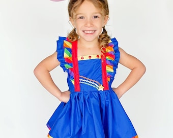 May include: A young girl wearing a blue dress with rainbow trim and a pink headband with colorful stars. She is smiling and has her hands on her hips.