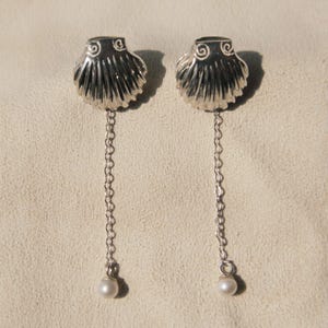 Könnte beinhalten: Ein Paar silberne Ohrringe mit Muschel-Design und einer baumelnden Perle am Ende jedes Ohrrings.