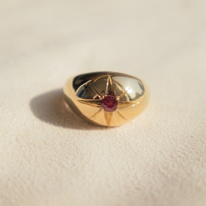 Op de afbeelding: Een goudkleurige ring met een sterrenpatroon en een kleine, ronde, rode edelsteen in het midden. De ring heeft een glad, gepolijst oppervlak en ligt op een crèmekleurige stof.
