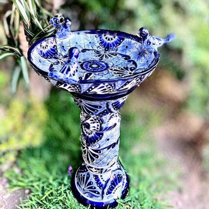 Pode incluir: Banho de p&aacute;ssaros de cer&acirc;mica azul e branco com tr&ecirc;s pequenos p&aacute;ssaros azuis empoleirados na borda. O banho de p&aacute;ssaros tem uma base de pedestal com intrincados desenhos florais.