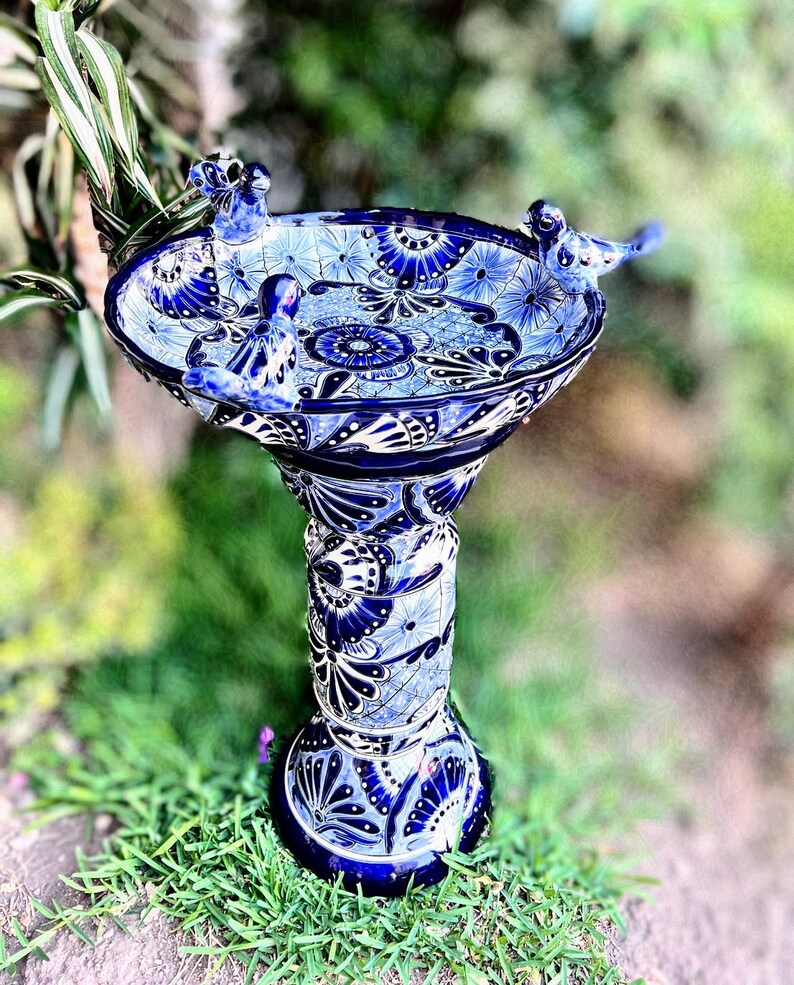 Pode incluir: Banho de p&aacute;ssaros de cer&acirc;mica azul e branco com tr&ecirc;s pequenos p&aacute;ssaros azuis empoleirados na borda. O banho de p&aacute;ssaros tem uma base de pedestal com intrincados desenhos florais.