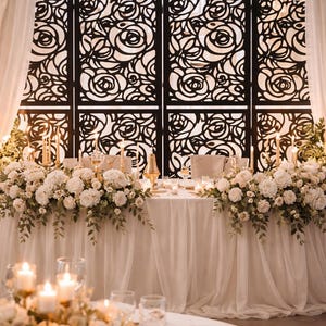 Puede incluir: Elegante decoración de recepción de bodas con una mesa adornada con rosas blancas y velas. Una pantalla decorativa con un diseño de rosas sirve como telón de fondo. Cortinas blancas suaves y arreglos florales crean un ambiente romántico.