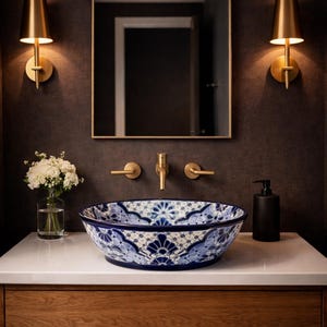 May include: A bathroom sink with a blue and white floral patterned ceramic bowl. The sink is set on a white countertop with a wooden cabinet. Gold fixtures and sconces complement the design. A mirror hangs above the sink.